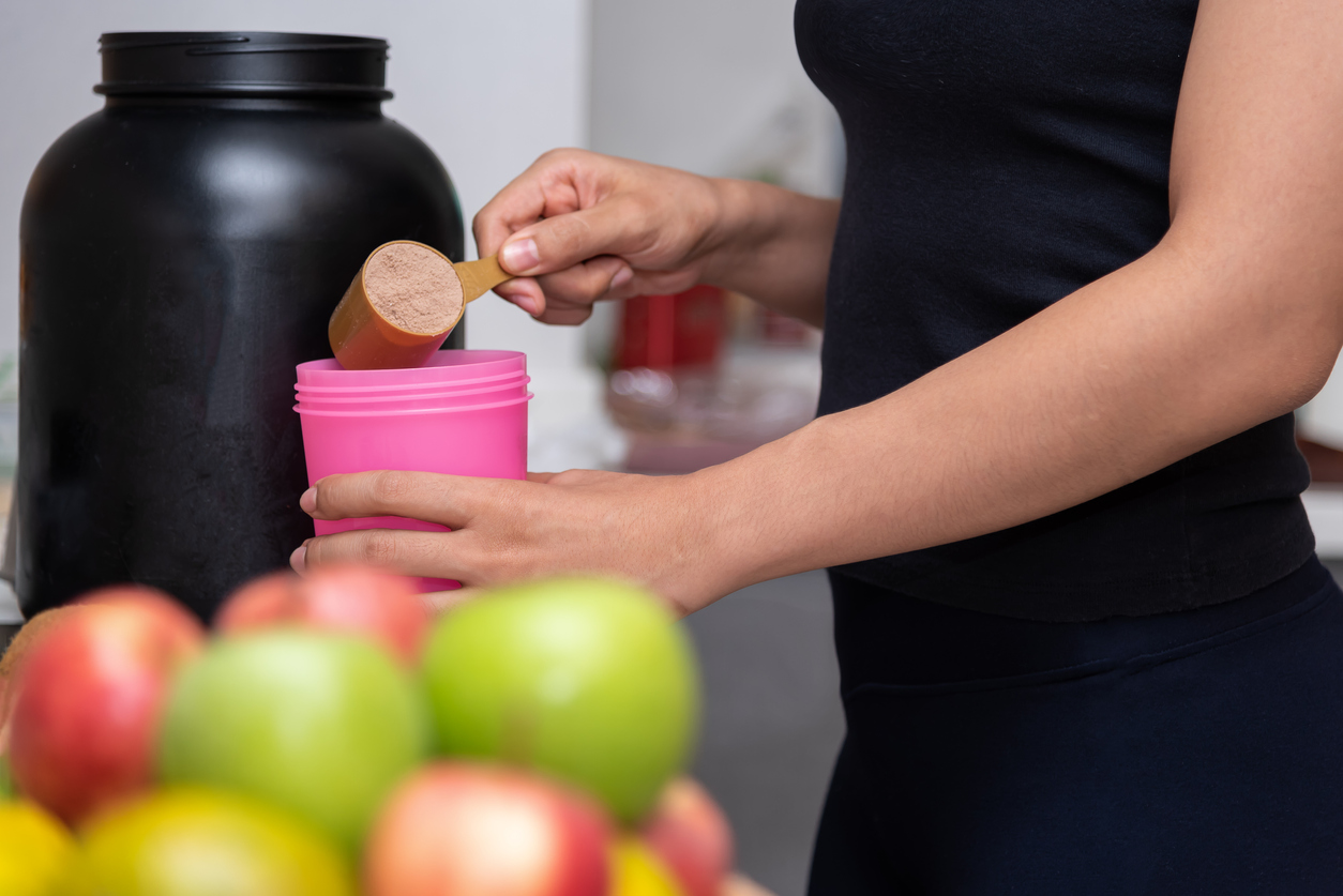 Healthy women preparing a powdered beverage whey protein after doing weight training in the kitchen with fresh fruits as a blurred foreground.